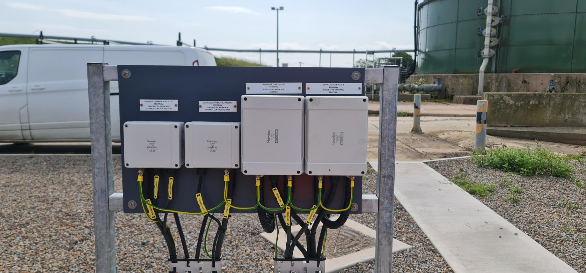 Industrial control boxes with wiring mounted on metal frame beside pipeline and gravel yard.