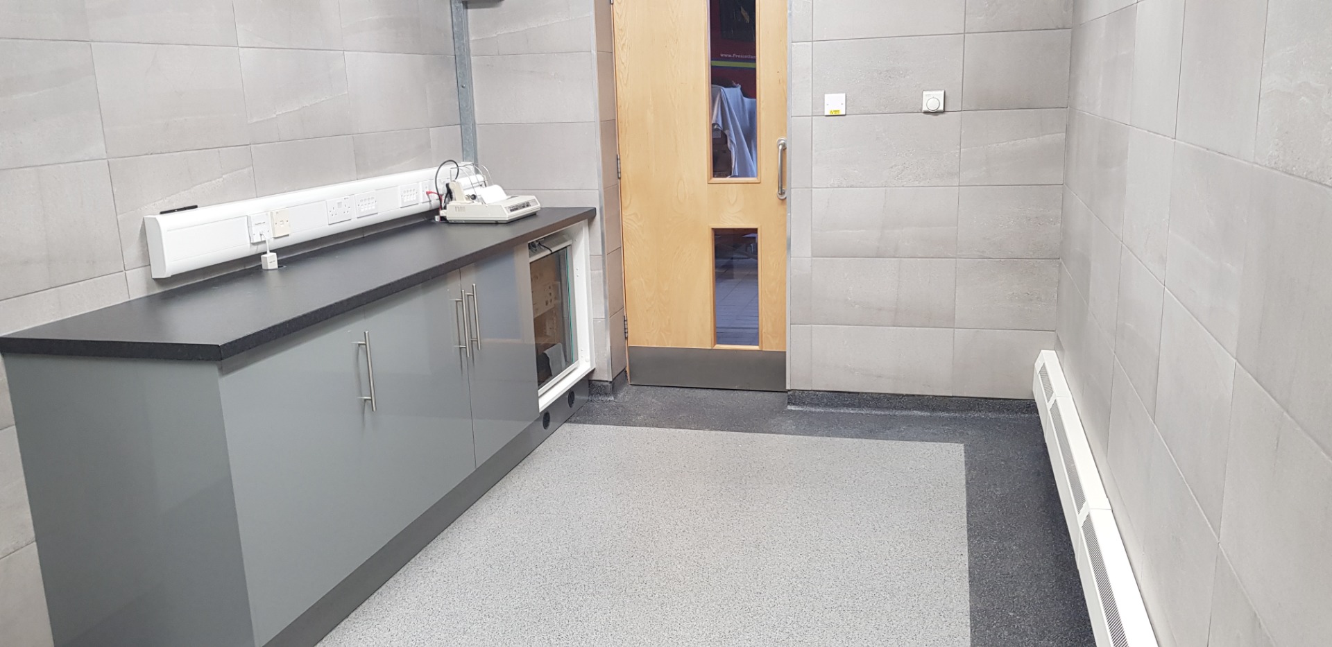 Clean utility room with gray countertops, built-in sink, medical equipment, and wooden door opening to hallway.