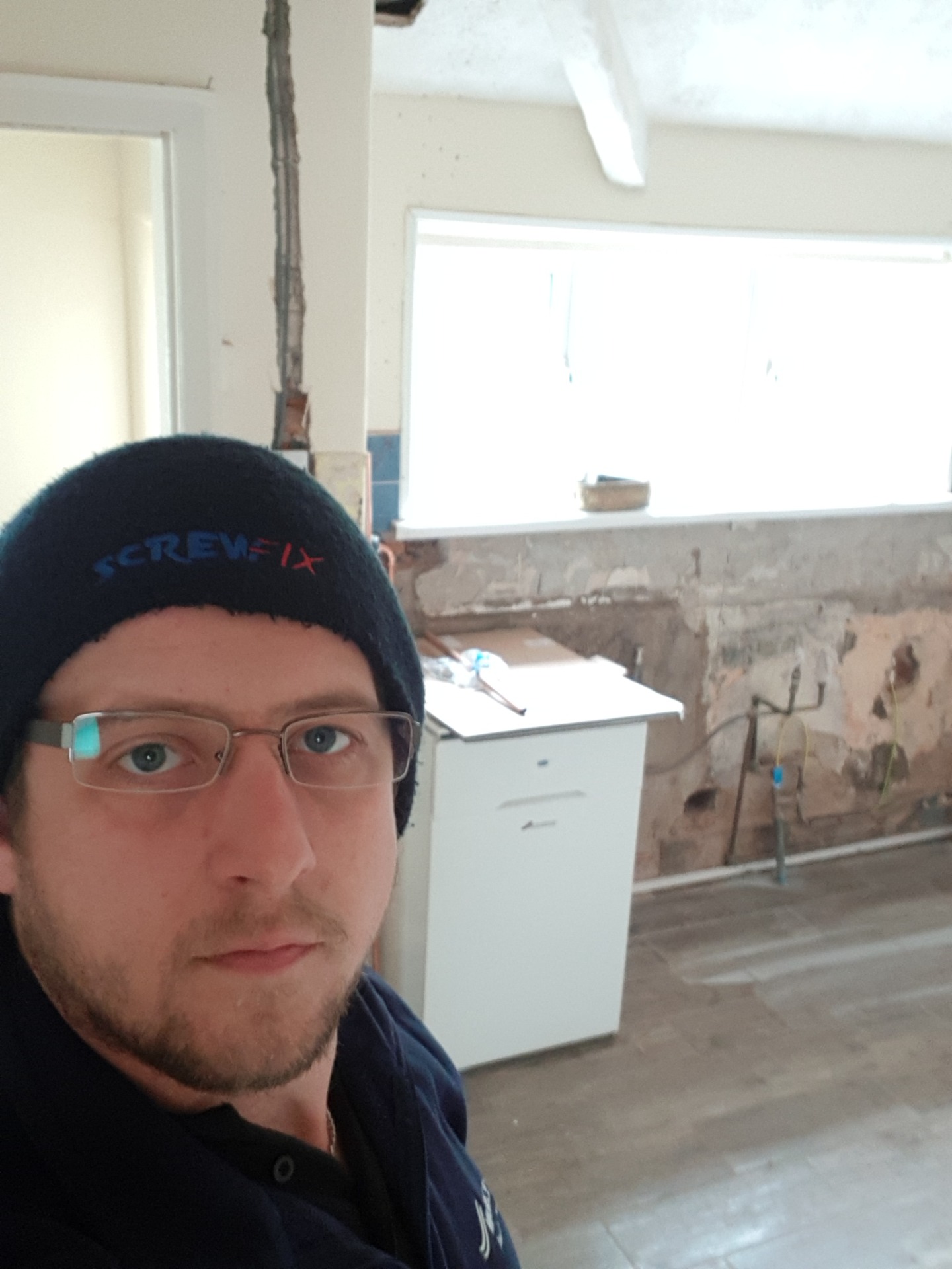 Man wearing glasses and beanie in a kitchen, with unfinished wall and tools in background.