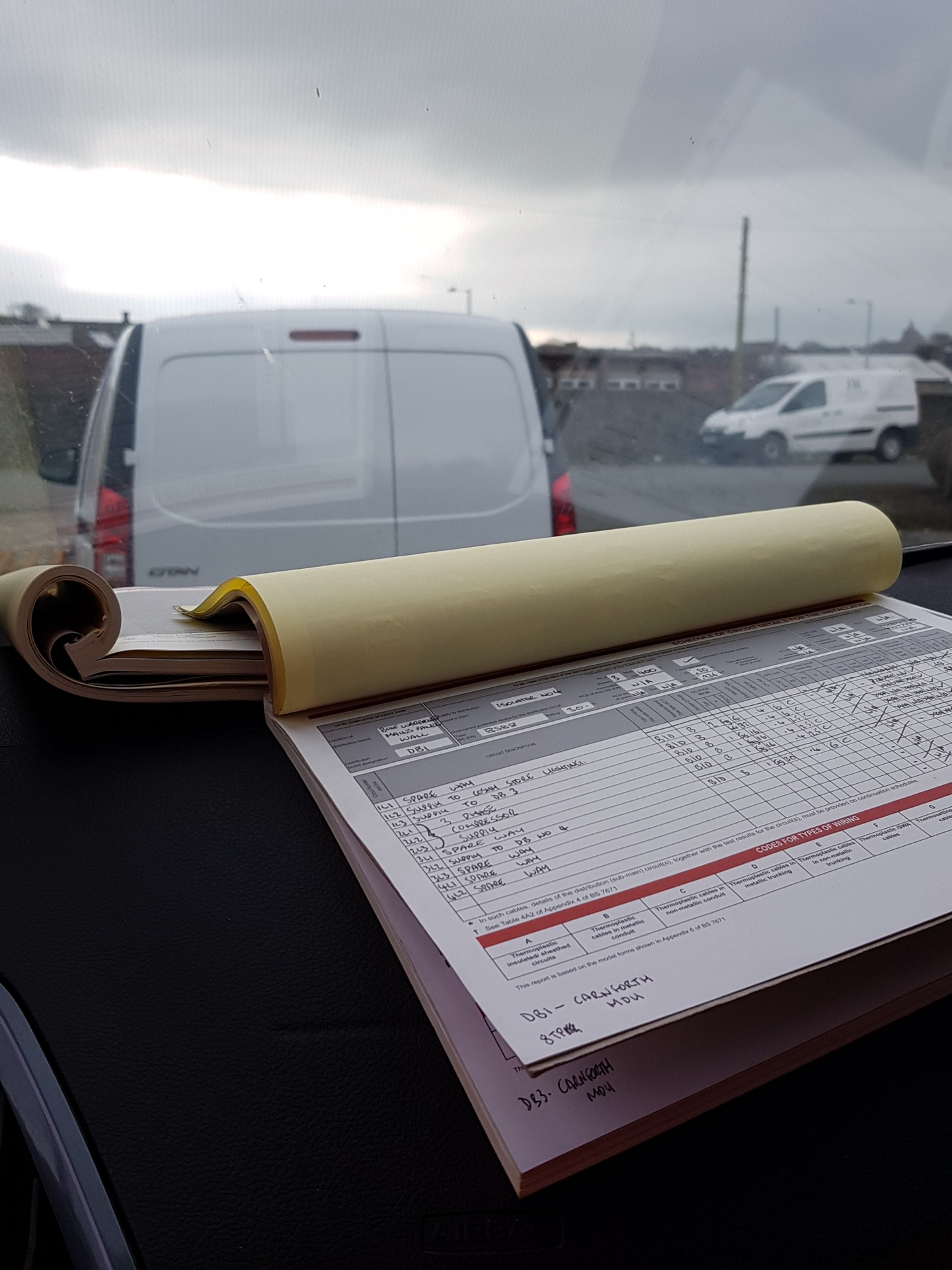 Open vehicle paperwork with handwritten notes and rolled plans on dashboard, viewed through windshield at parking lot.